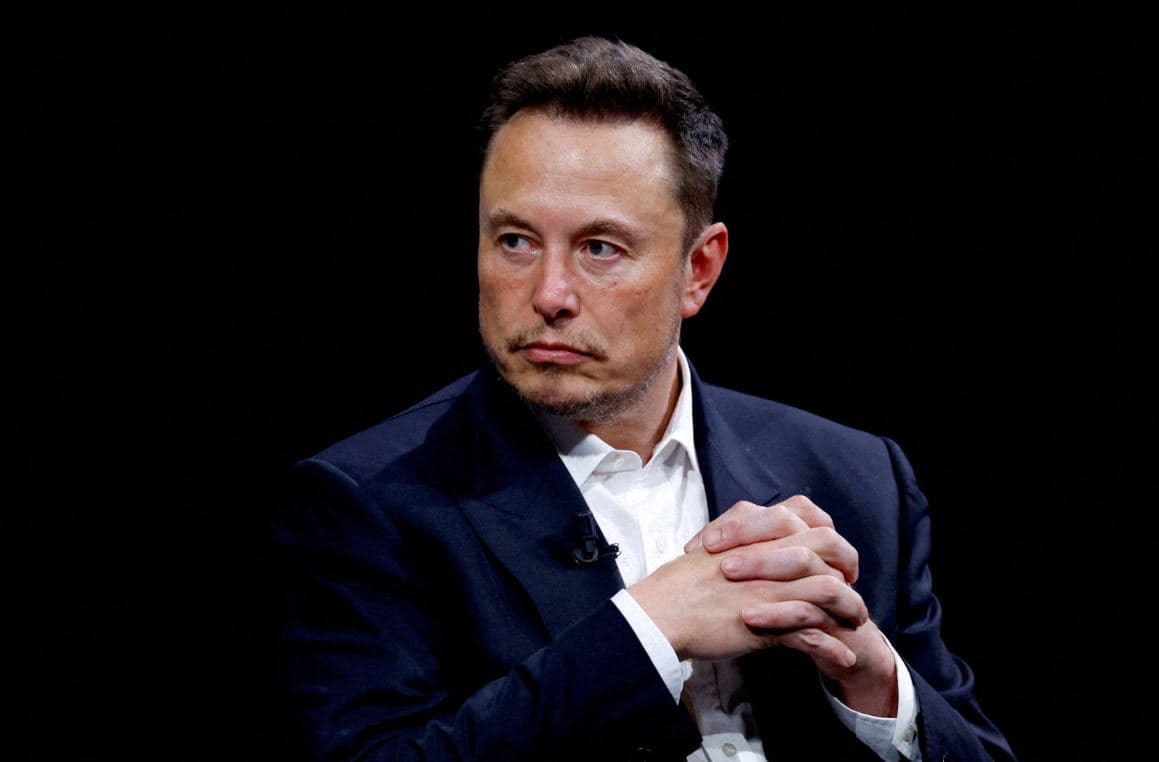 Elon Musk in a formal black suit, seated with hands clasped, looking serious during a public appearance or interview.