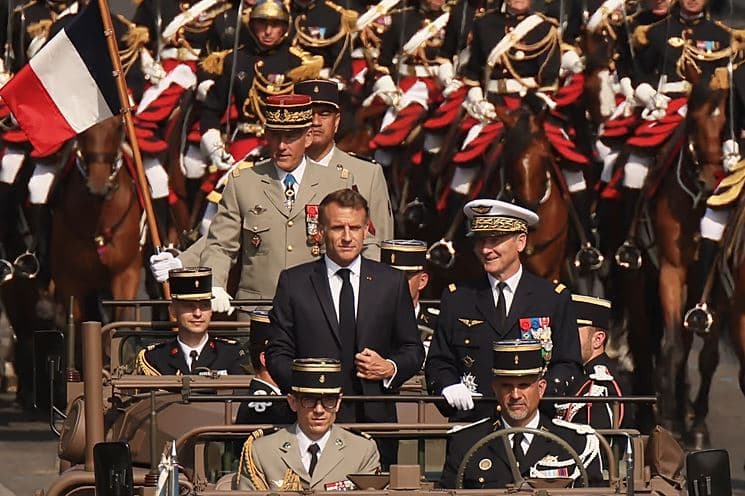 French President Emmanuel Macron rides in a military vehicle alongside senior officers during a ceremonial parade, with mounted cavalry and French flags visible in the background.