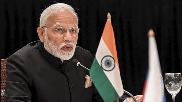 Narendra Modi speaking at an official event with the Indian flag in the foreground.