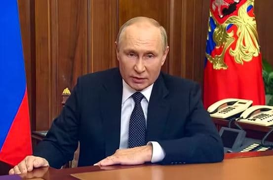 Vladimir Putin delivering a televised national address seated at the Kremlin desk with the Russian flag and state coat of arms visible behind him